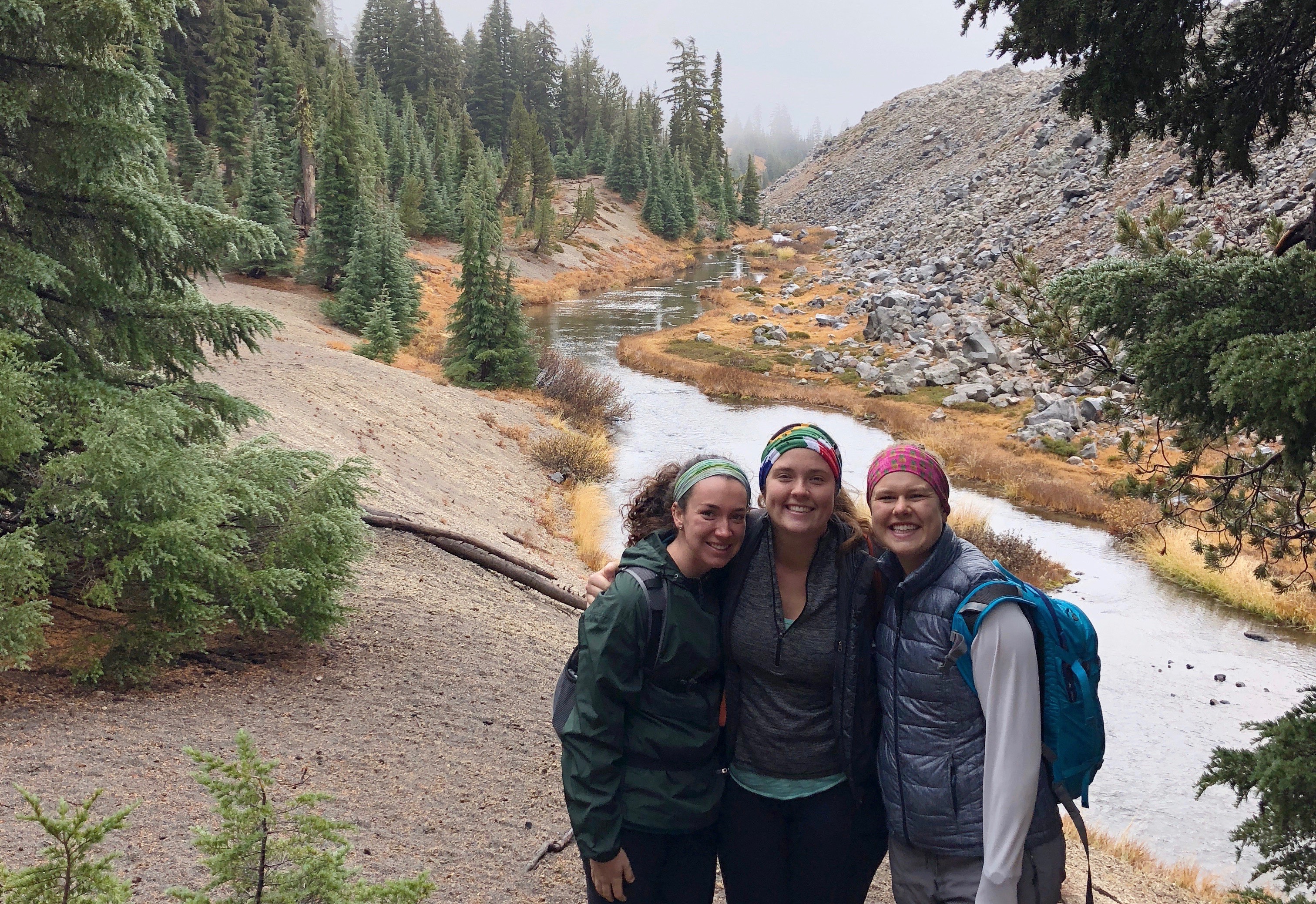 Three people standing close together by a river with pine trees