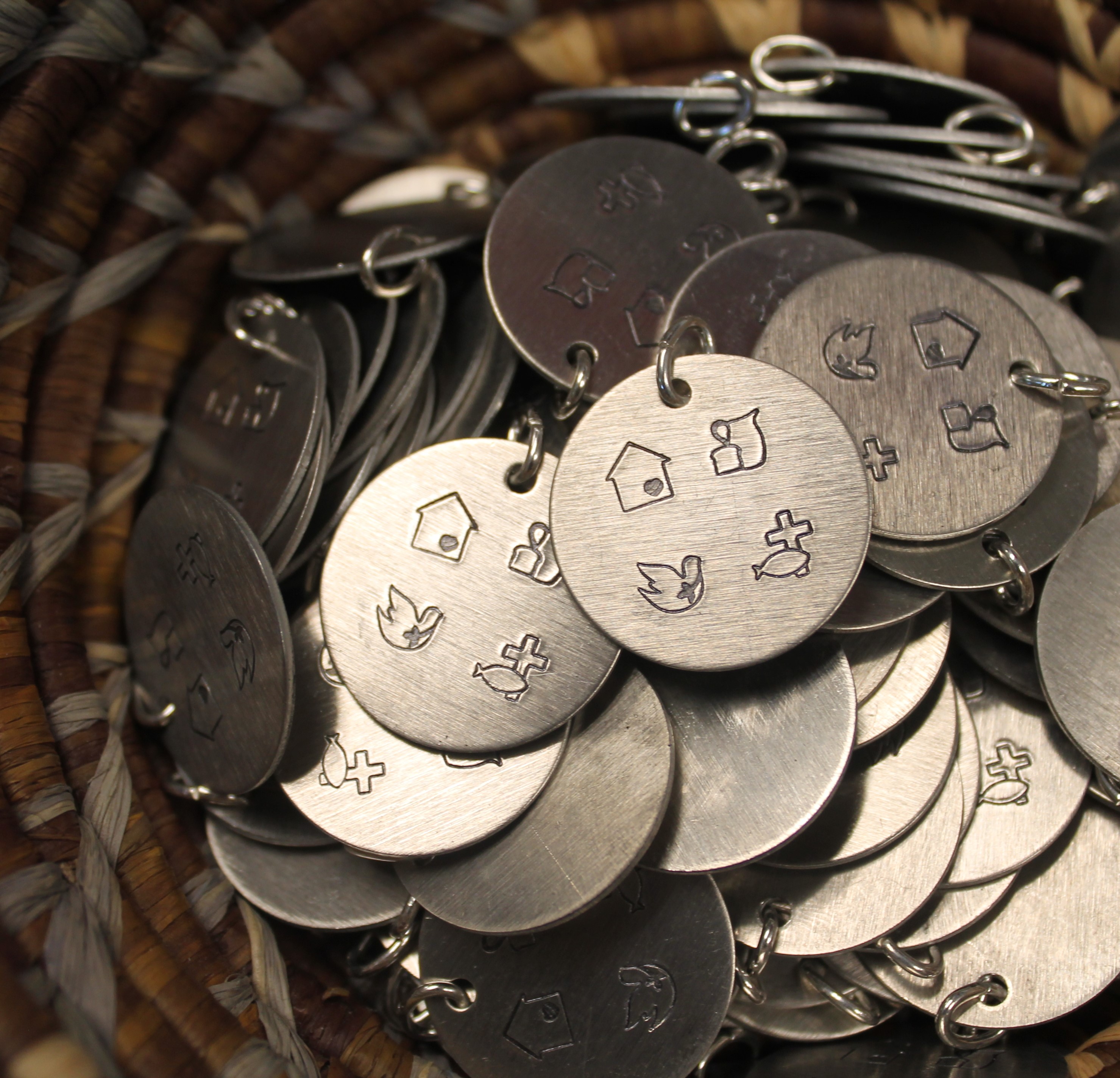 round stainless steel pendants in a basket. Each pendant has an etched stamp of a house, bread and a jug, a dove, and a fish and cross