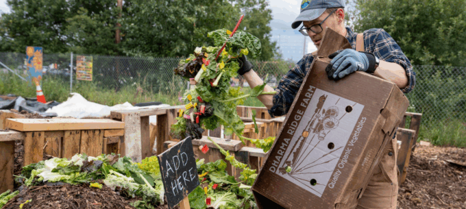 Jesuit Volunteer in hat and glasses emptying a box of compost into a garden compost pile during service