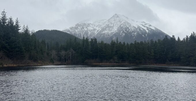 Snowy mountain in the background, with a lake in the foreground, evergreen trees in between. Overcast day.