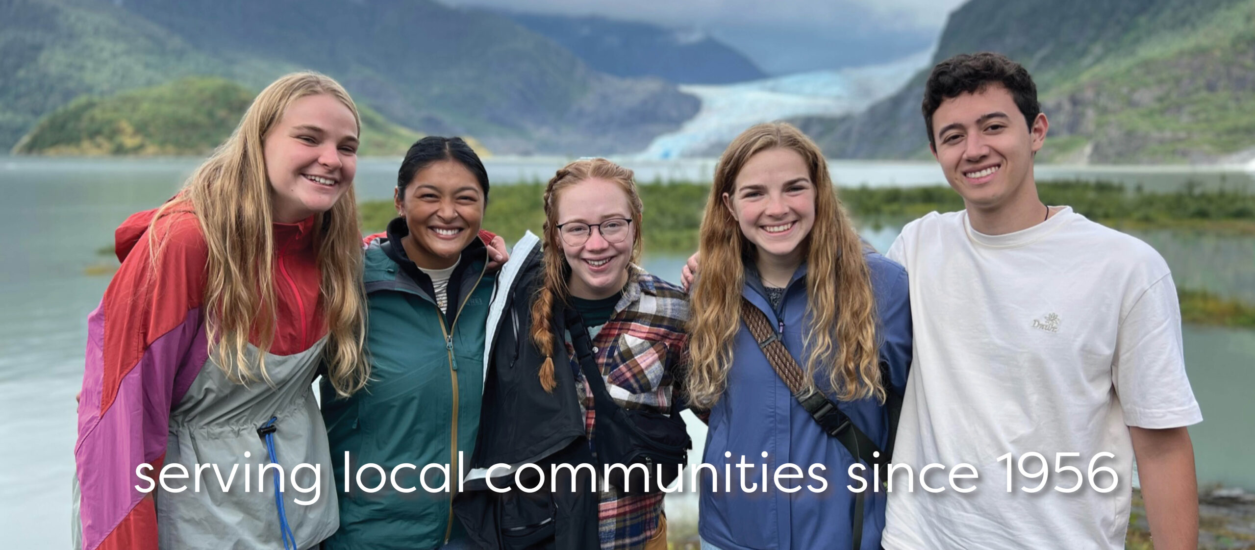 five volunteers smiling in front of a glacier with the words "serving local communities since 1956"